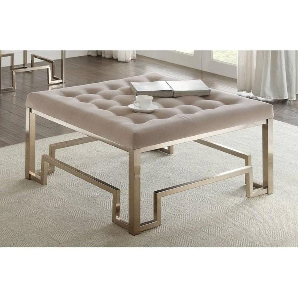 Square champagne polyester tufted cocktail ottoman with a gold metal geometric base on a light rug.