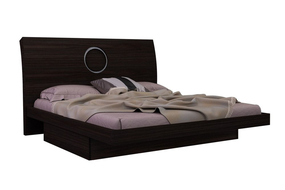 Dark brown wood platform bed with a circular accent on the headboard, dressed with purple sheets and a taupe blanket.