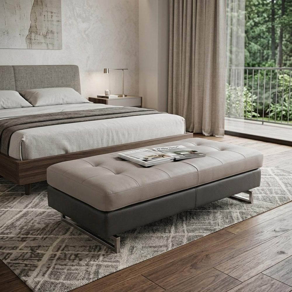 63" Taupe Brown and Silver Faux Leather Tufted Ottoman styled in a furnished interior with a light gray wall on a dark wood floor.