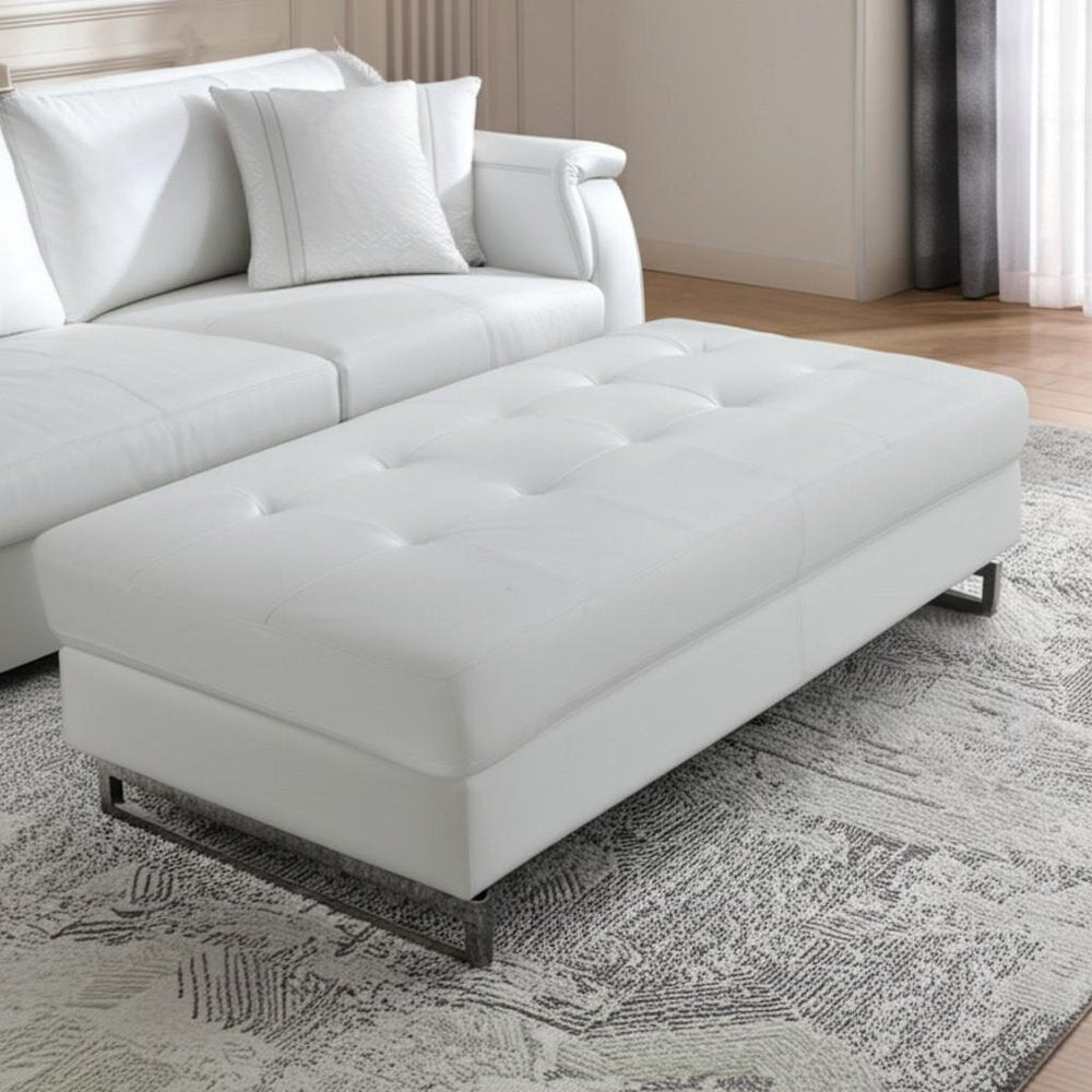 63" White and Silver Faux Leather Ottoman styled in a furnished interior with a tan wall on a tiled floor.