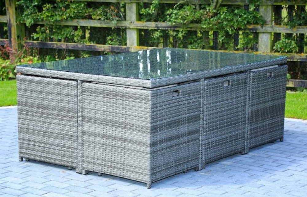 Gray rattan dining set with glass top on a paved patio with a wooden fence and greenery in the background.
