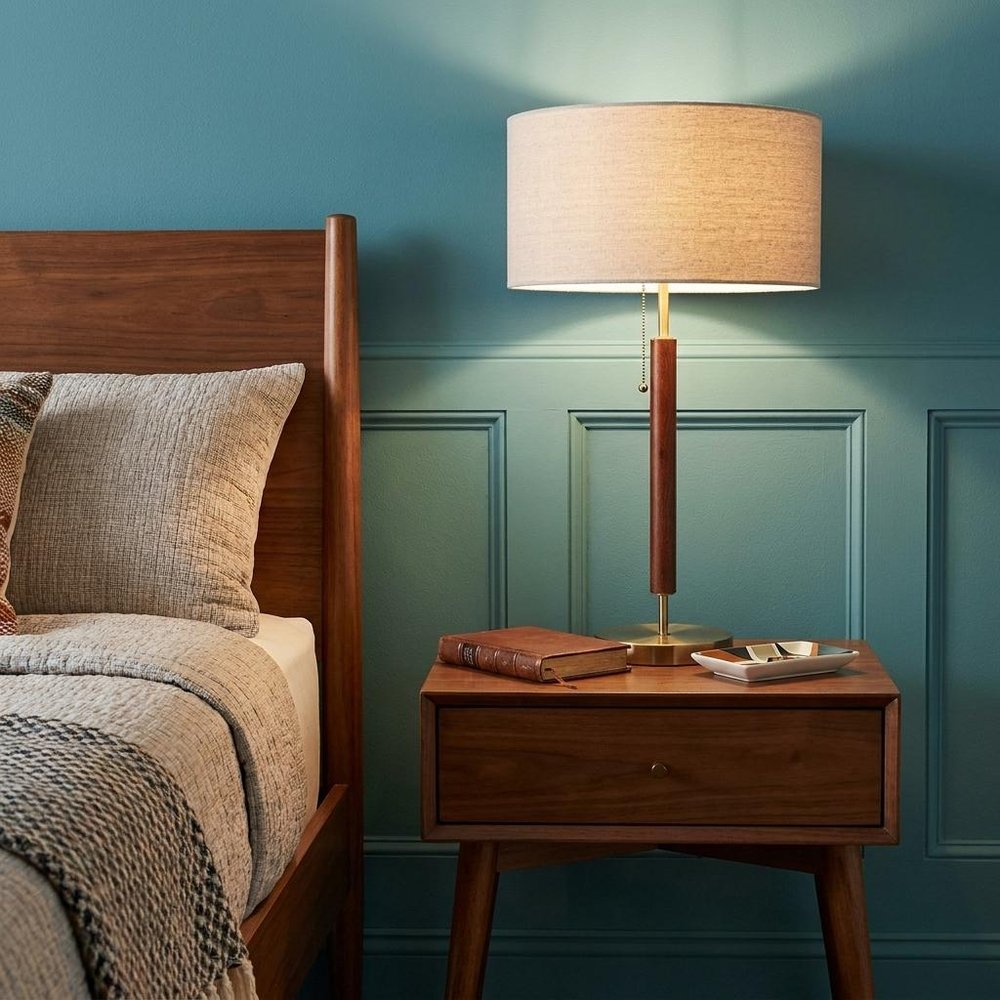 26" Antiqued Brass Solid Wood Table Lamp With Beige Drum Shade shown outdoors on an outdoor setting with greenery.