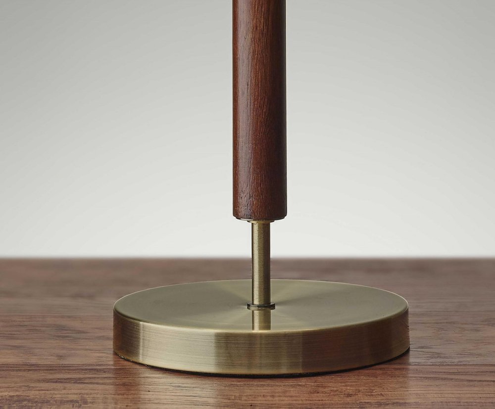 26" Antiqued Brass Solid Wood Table Lamp With Beige Drum Shade displayed on a dark wood tabletop in a furnished interior against a light gray wall.