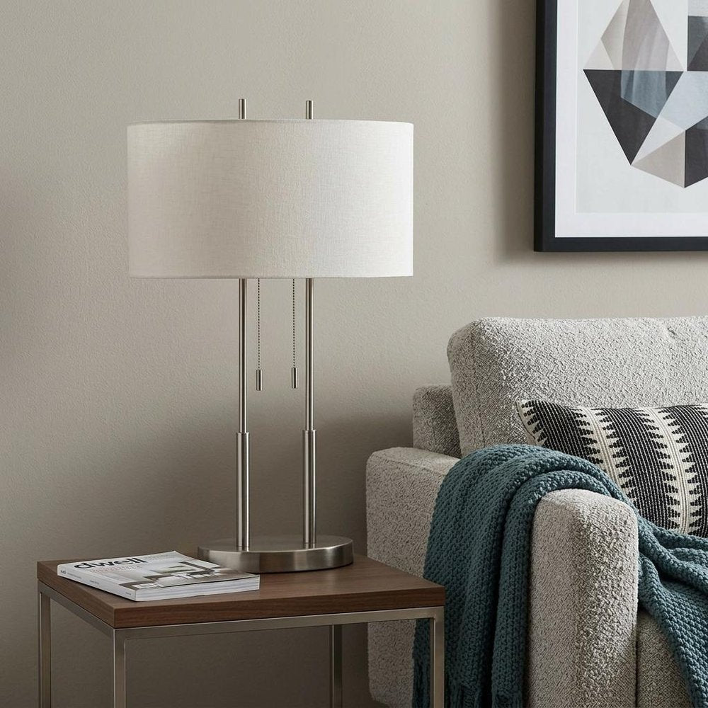 27" Silver Metal Two Light Bedside Lamp With Off White Drum Shade styled in a furnished interior with a light gray wall on a dark wood floor in off-white metal.