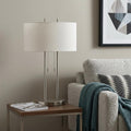 27" Silver Metal Two Light Bedside Lamp With Off White Drum Shade styled in a furnished interior with a light gray wall on a dark wood floor in off-white metal.