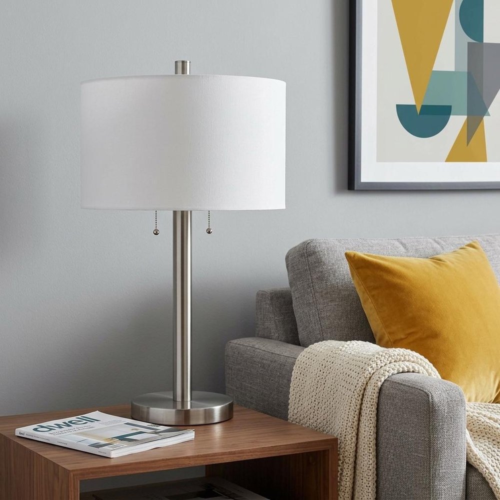 28" Silver Metal Two Light Bedside Table Lamp With White Drum Shade displayed on a dark wood tabletop in a furnished interior against a light gray wall.