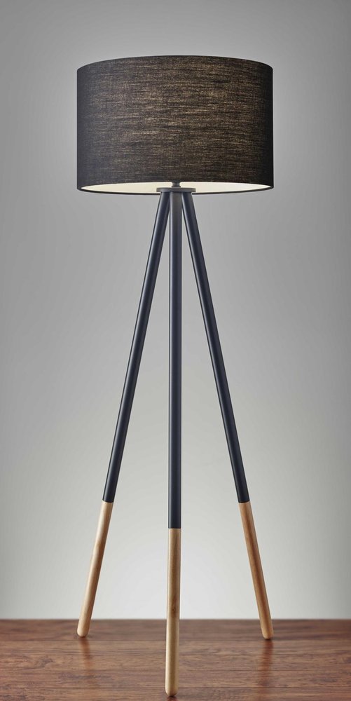 Black metal tripod floor lamp with a black fabric drum shade and wood accents, set against a grey background and wooden floor.