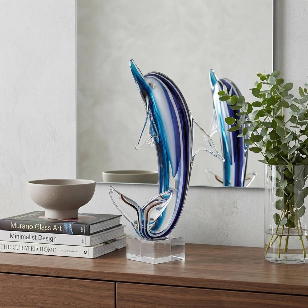 14" Blue Murano Glass Dolphin Figurine Tabletop Sculpture styled in a furnished interior with an off-white wall on a dark wood floor.