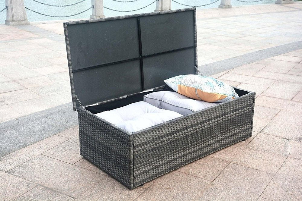 Gray wicker outdoor storage chest with lid open, showing white and patterned cushions on a stone patio.