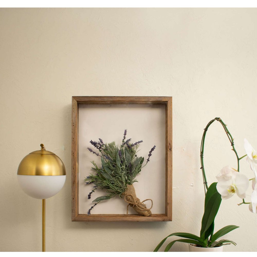 A framed bouquet of lavender and sage with burlap tie on a cream wall next to a gold and white lamp and a white orchid.
