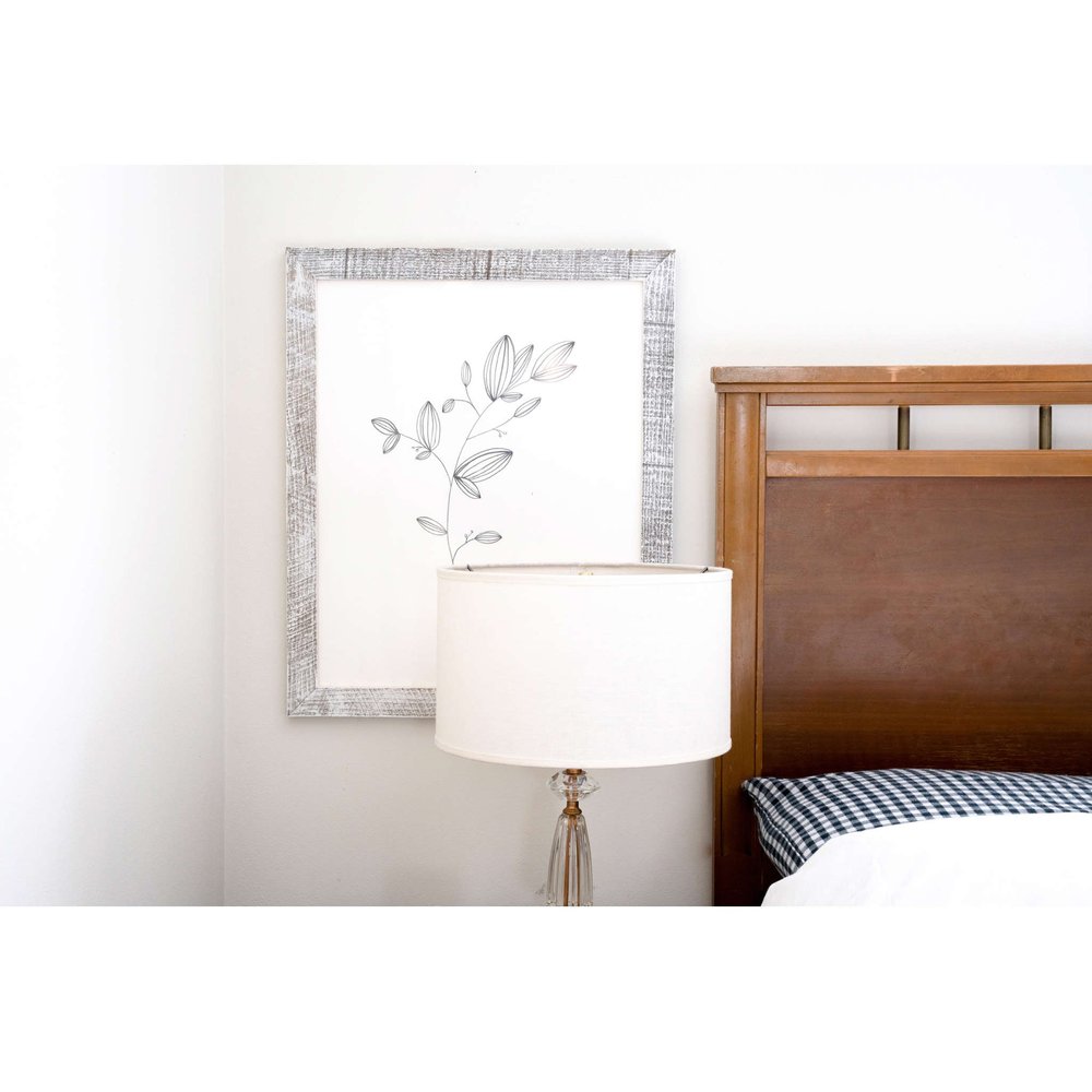 White solid wood tabletop picture frame with a botanical line drawing, next to a bedside lamp and wooden headboard.