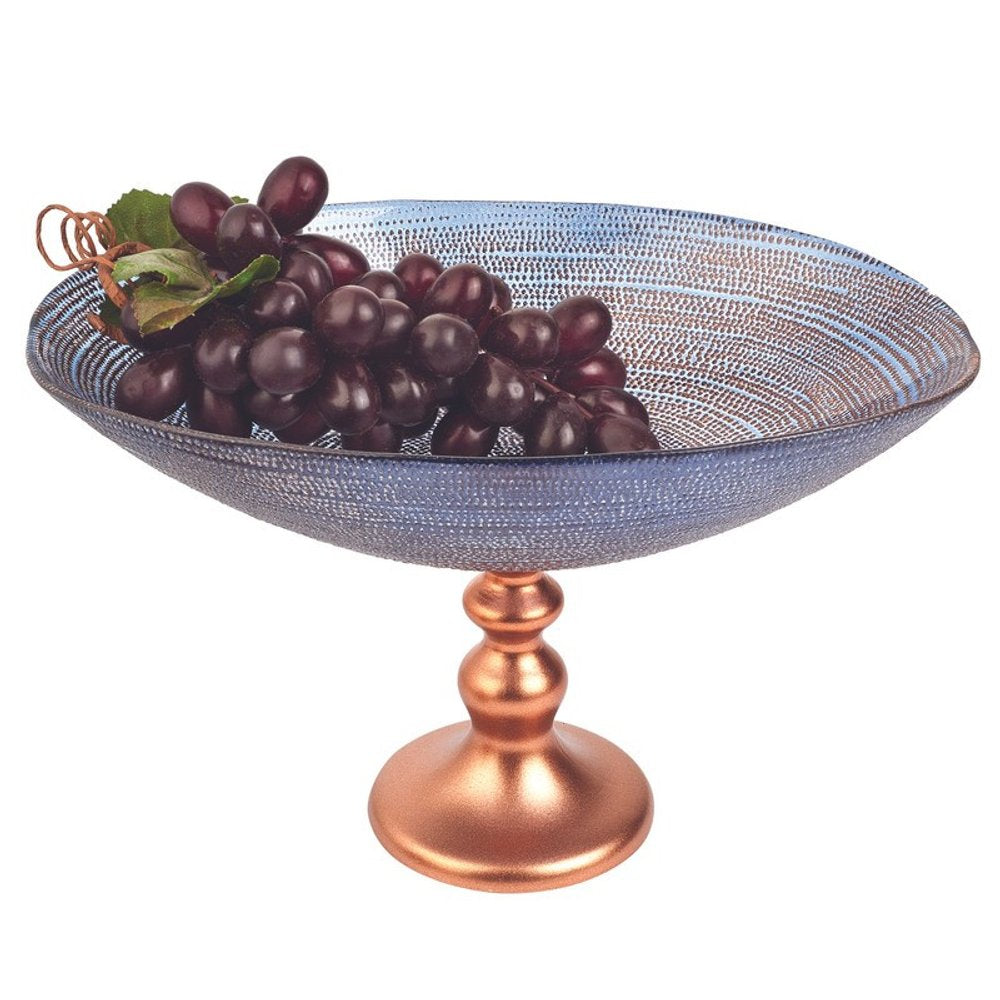 A blue glass footed bowl with a copper base holds dark red grapes and green leaves against a white background.