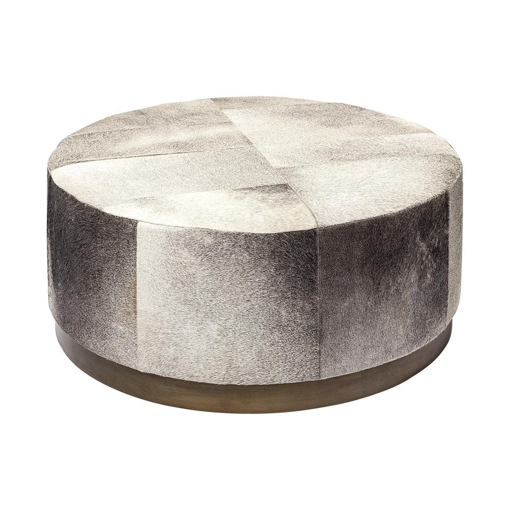 Round ottoman with patchwork gray and white cowhide upholstery and a brass base on a white background.