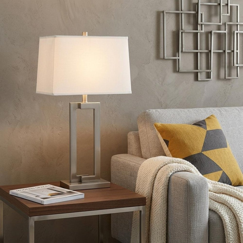 30" Silver Metal Table Lamp With White Rectangular Shade displayed on a dark wood tabletop in a furnished interior against a tan wall.