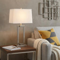 30" Silver Metal Table Lamp With White Rectangular Shade displayed on a dark wood tabletop in a furnished interior against a tan wall.