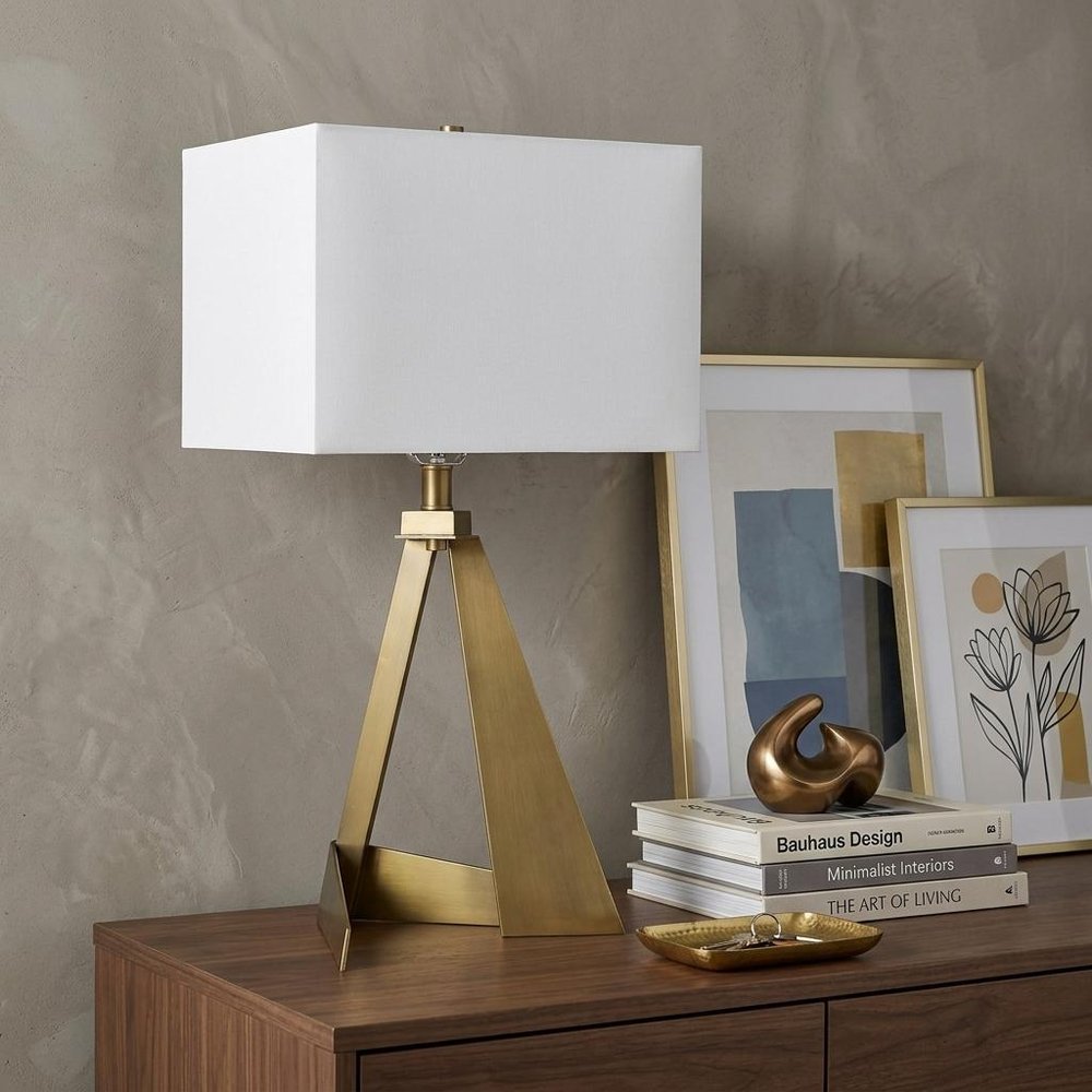 25" Brass Novelty Table Lamp With Shade displayed on a dark wood tabletop in a furnished interior against a tan wall.