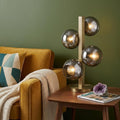 24" Brass Metal Four Light Novelty Table Lamp With Silver Metallic Globe Shades displayed on a dark wood tabletop in a furnished interior against a olive wall.