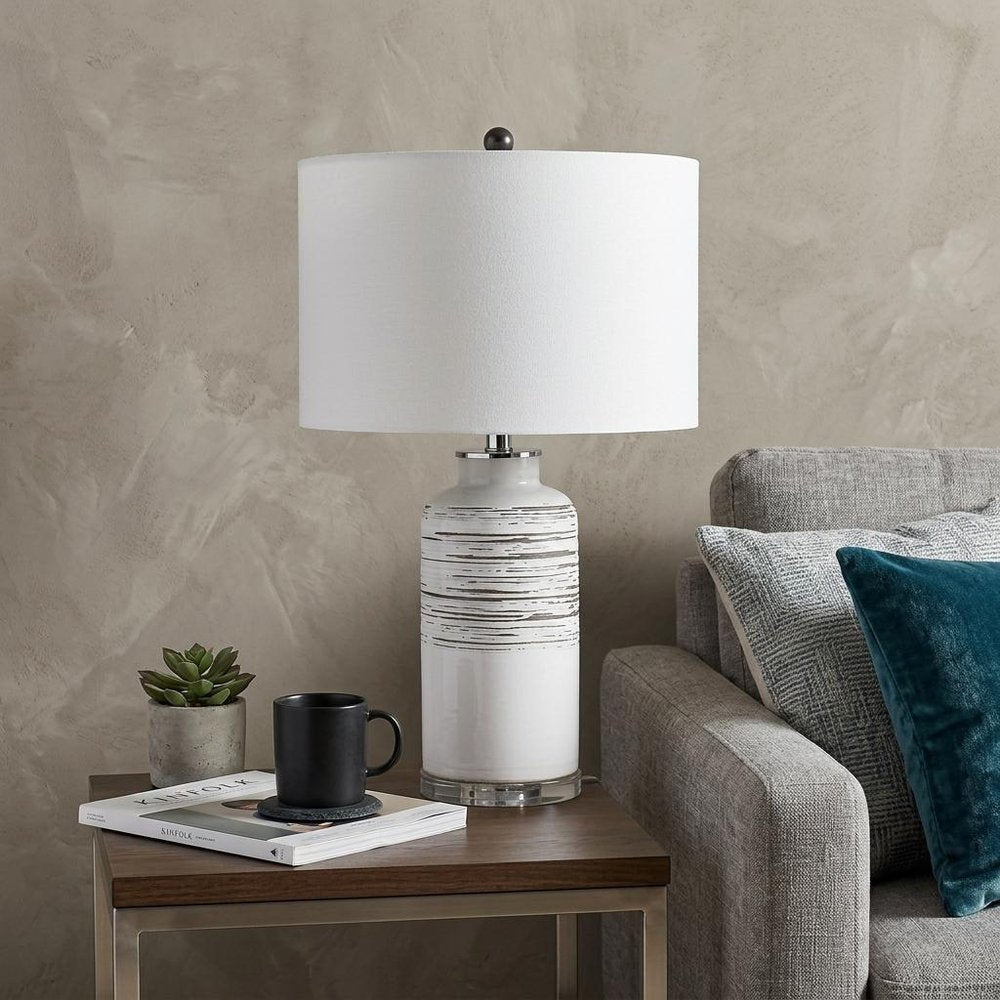25" Black and White Ceramic Column Lamp With White Drum Shade styled in a furnished interior with a tan wall on a dark wood floor.