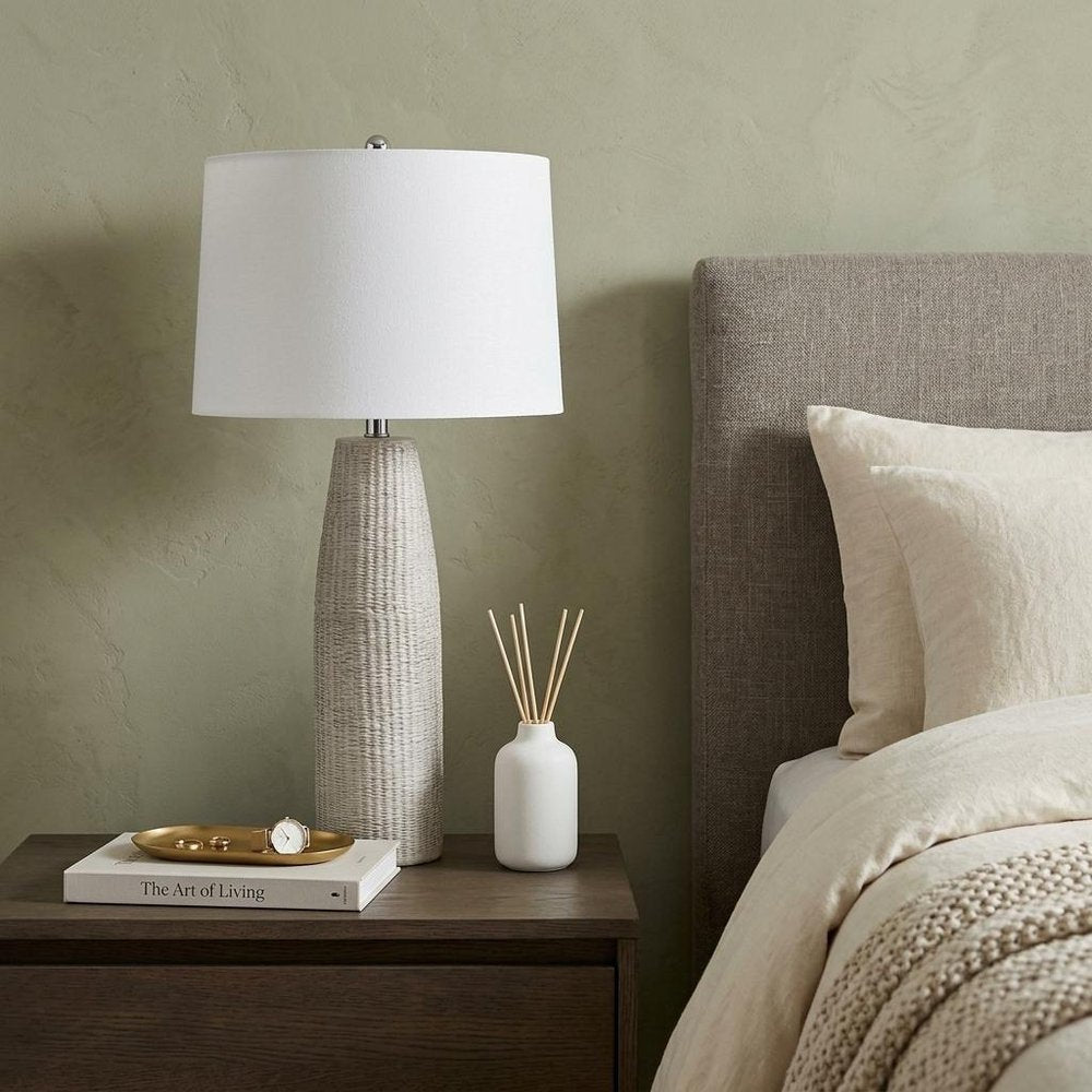 33" Light Gray Ceramic Table Lamp With White Drum Shade displayed on a dark wood tabletop in a furnished interior against a gold wall.