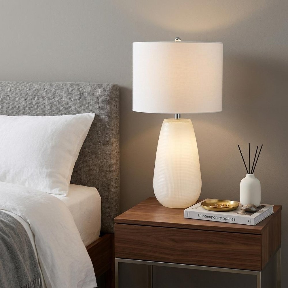 28" White Glass Table Lamp With White Drum Shade displayed on a dark wood tabletop in a furnished interior against a tan wall.
