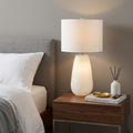 28" White Glass Table Lamp With White Drum Shade displayed on a dark wood tabletop in a furnished interior against a tan wall.