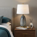 30" Teal Blue Concrete Table Lamp With Cream Empire Shade displayed on a dark wood tabletop in a furnished interior against a tan wall.