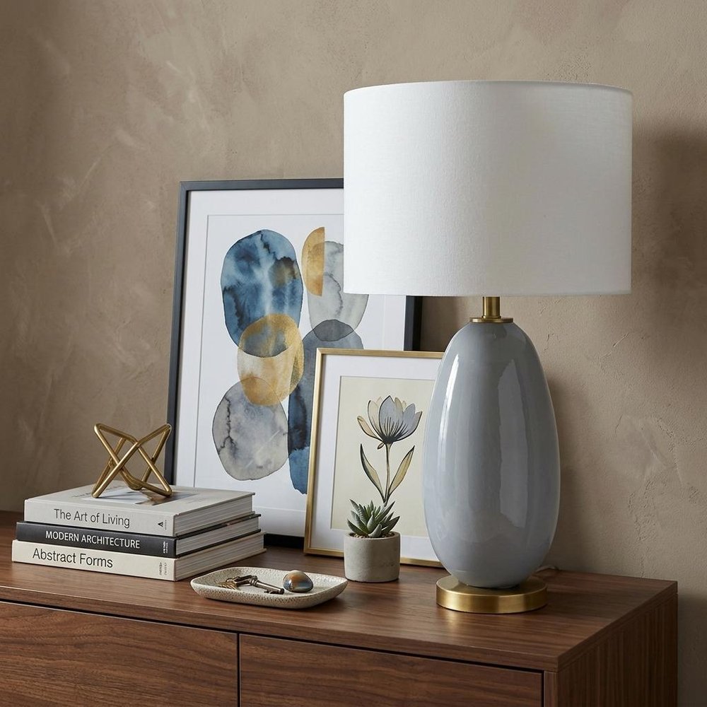 29" Light Gray Glass Table Lamp With White Drum Shade displayed on a dark wood tabletop in a furnished interior against a tan wall.