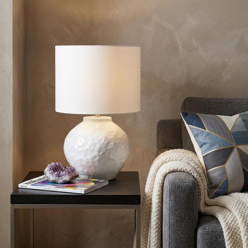 26" White Ceramic Table Lamp With White Drum Shade displayed on a dark wood tabletop in a furnished interior against a tan wall.