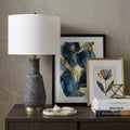 26" Distressed Black and Gold Ceramic Table Lamp With White Drum Shade displayed on a dark wood tabletop in a furnished interior against a gray wall.