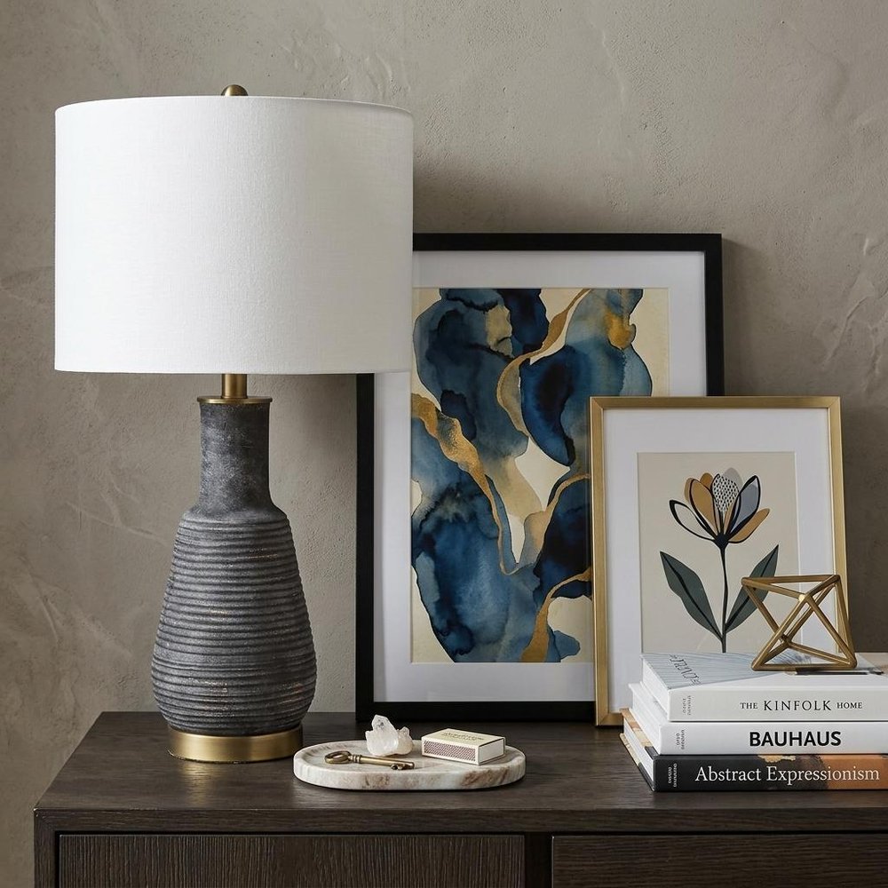 26" Distressed Black and Gold Ceramic Table Lamp With White Drum Shade displayed on a dark wood tabletop in a furnished interior against a gray wall.