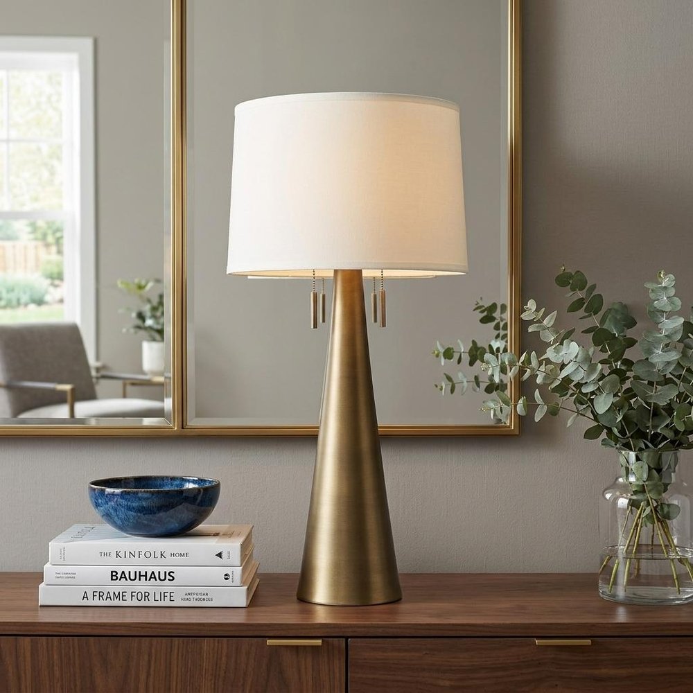 34" Brass Metal Two Light Table Lamp With White Empire Shade displayed on a dark wood tabletop in a furnished interior against a gray wall.