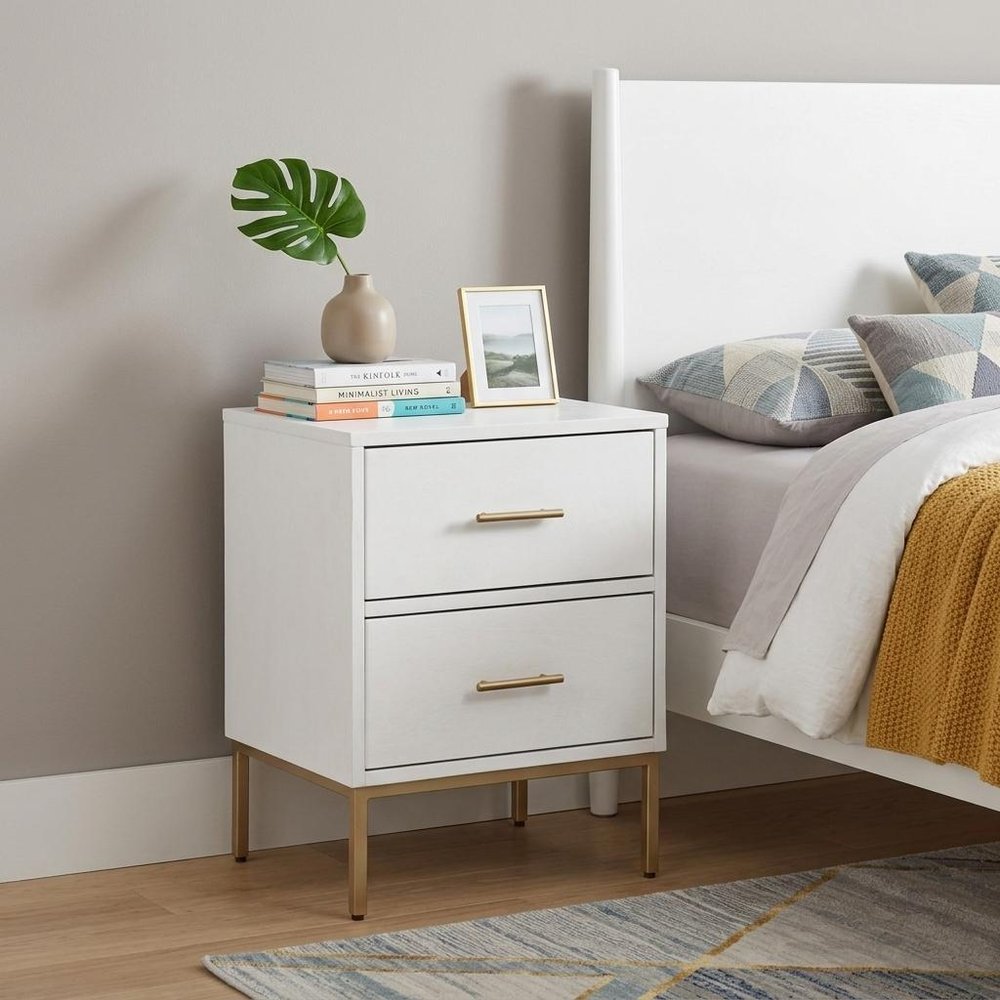 26" White And Gold Wood And Metal Two Drawer Nightstand styled in a furnished interior with a light gray wall on a dark wood floor.