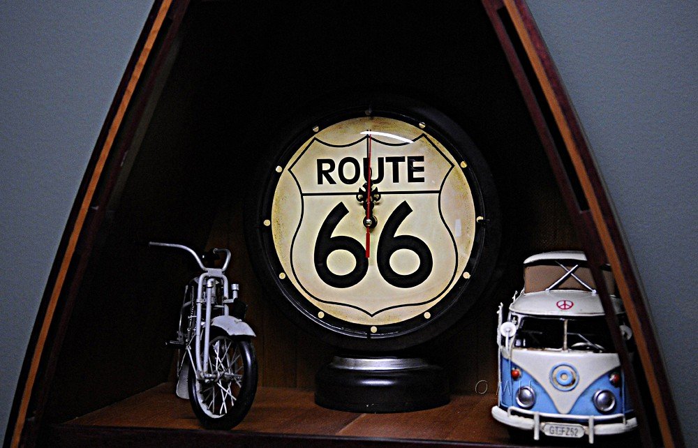 Black metal analog round wall clock with Route 66 design, featuring a miniature motorcycle and a blue and white camper van on a wooden shelf.