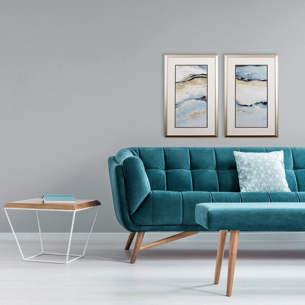 Two framed abstract wall art pieces with sandy beach colors hang above a teal velvet sofa and bench in a room with a gray wall.
