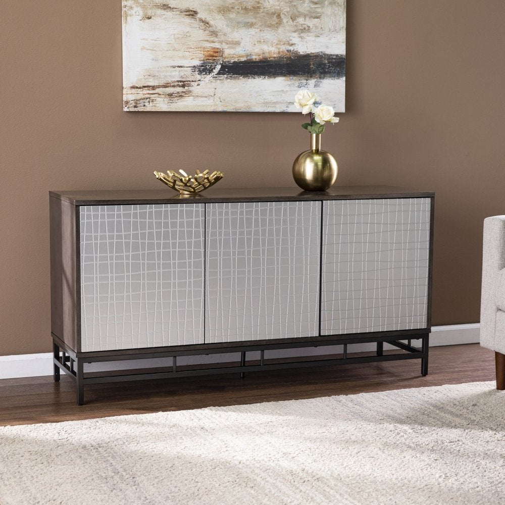A dark wood accent cabinet with three silver doors featuring a grid pattern sits on a dark metal base in front of a brown wall.