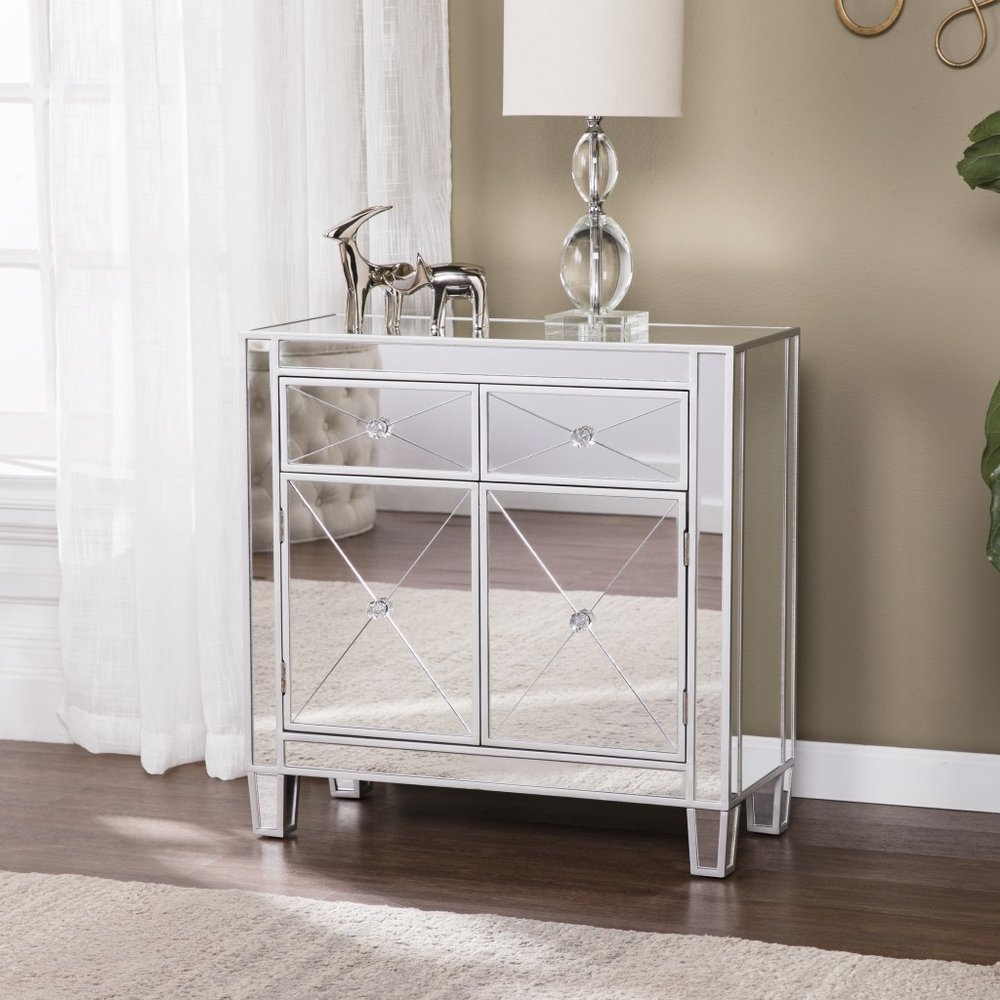 Mirrored accent cabinet with two doors and two drawers, featuring crystal accents, on a wooden floor with a sheer curtain in the background.