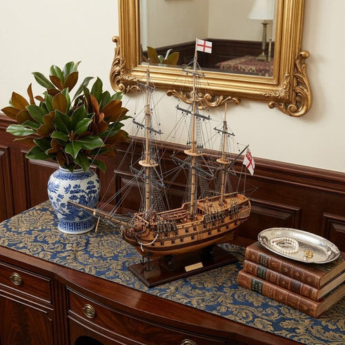 Wooden model ship with brown hull and white flags on a dark blue and gold patterned runner, placed on a wooden dresser.