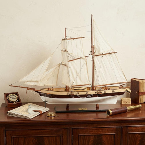 Model sailboat with white sails and brown hull on a wooden stand, with a compass, map, and spyglass on a wooden dresser.