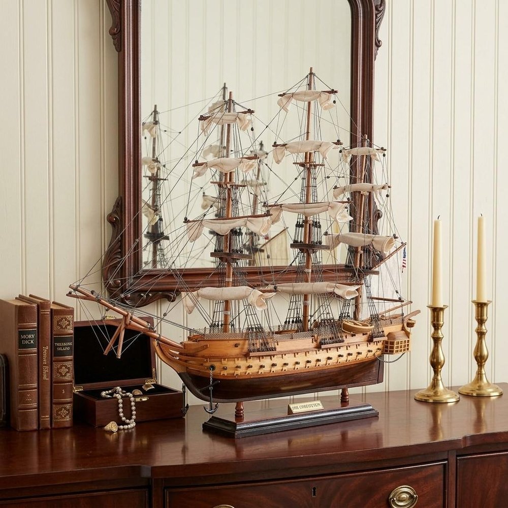 20" Wood Brown Solid Wood Hand Painted 1797 USS Constitution Small Model Boat styled in a furnished interior with a cream wall on a black floor.