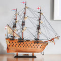 18" Wood Brown HMS Victory Hand Painted Decorative Boat styled in a furnished interior with an off-white wall on a off-white floor.