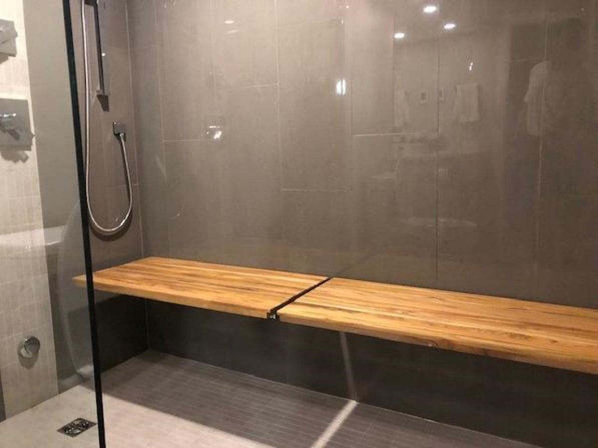 24" Premium Wall Mount Teak Shower Bench styled in a furnished interior with a tan wall on a dark wood floor in brown teak.