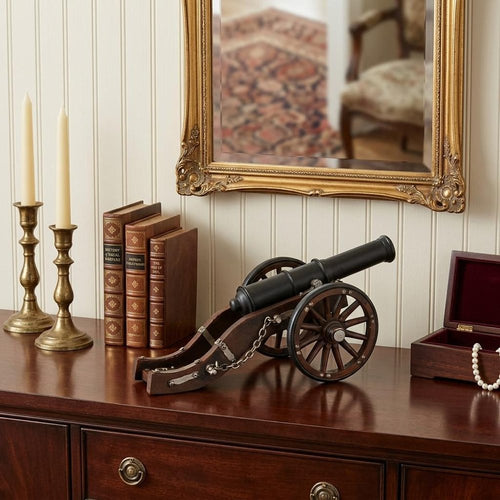 A decorative black and brown metal cannon sculpture with wooden wheels sits on a dark wood dresser, with books and candlesticks nearby.