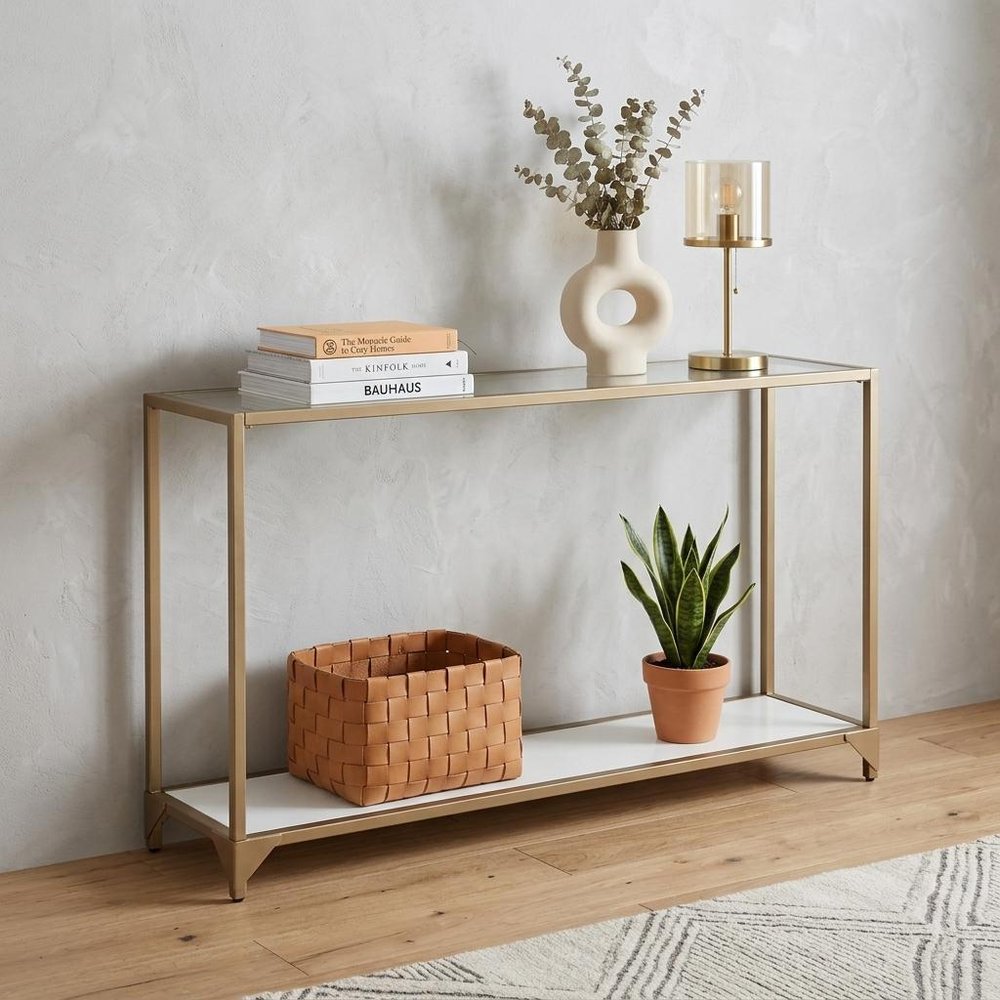 56" Clear and Gold Glass Mirrored Frame Console Table With Shelves styled in a furnished interior with an off-white wall on a wood floor.
