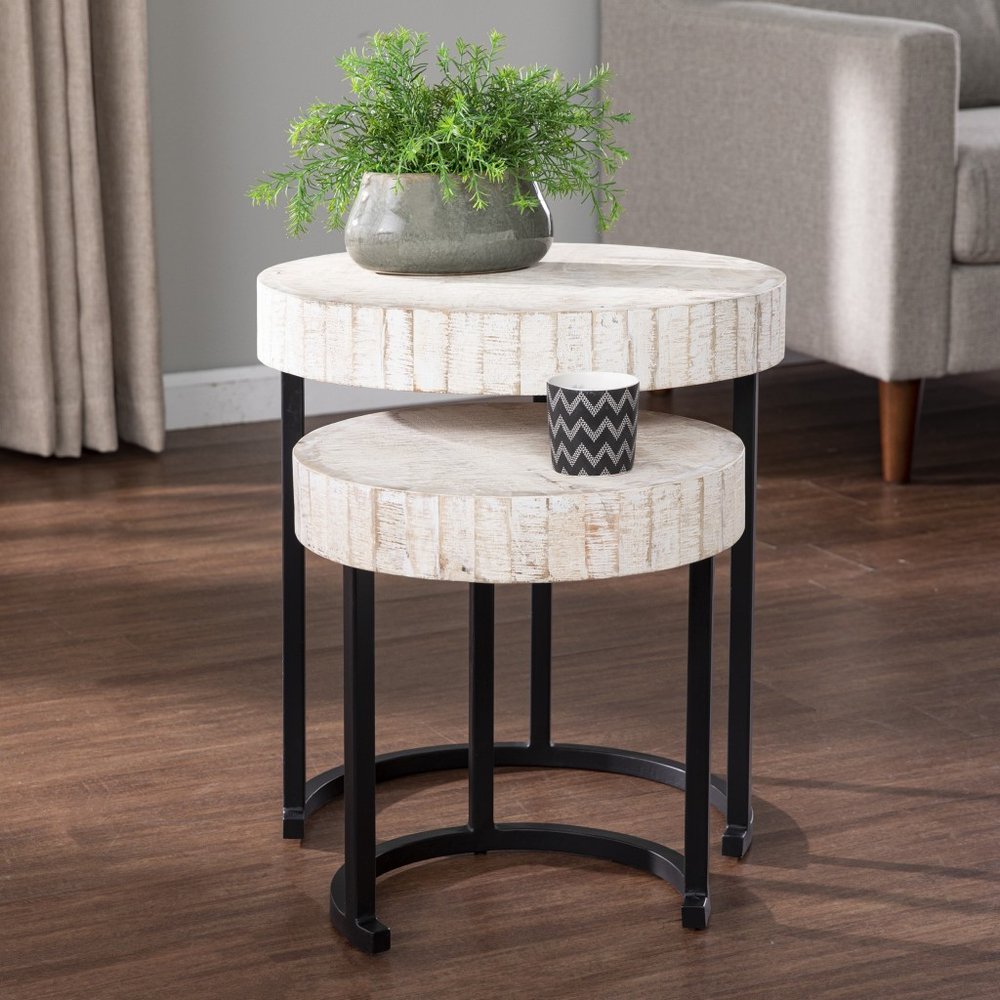 Two round white wood nested tables with black iron legs on a wooden floor, with a plant and candle on top.