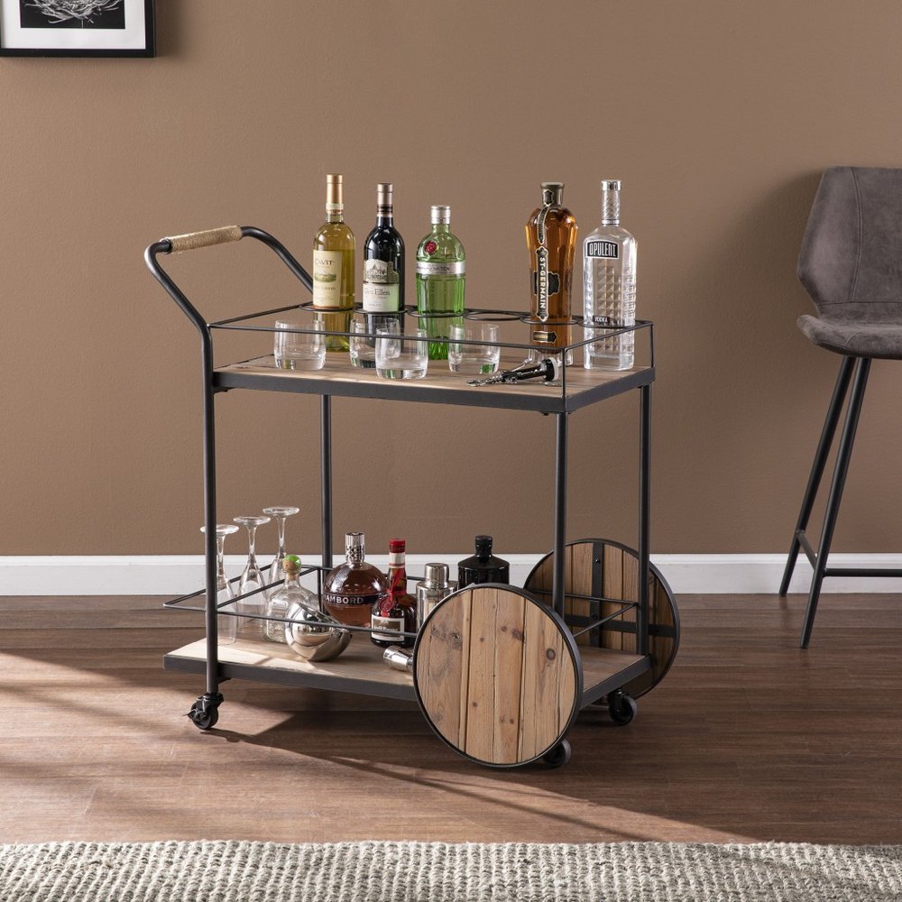 Black iron and reclaimed wood rolling bar cart with bottles and glasses on a wooden floor.