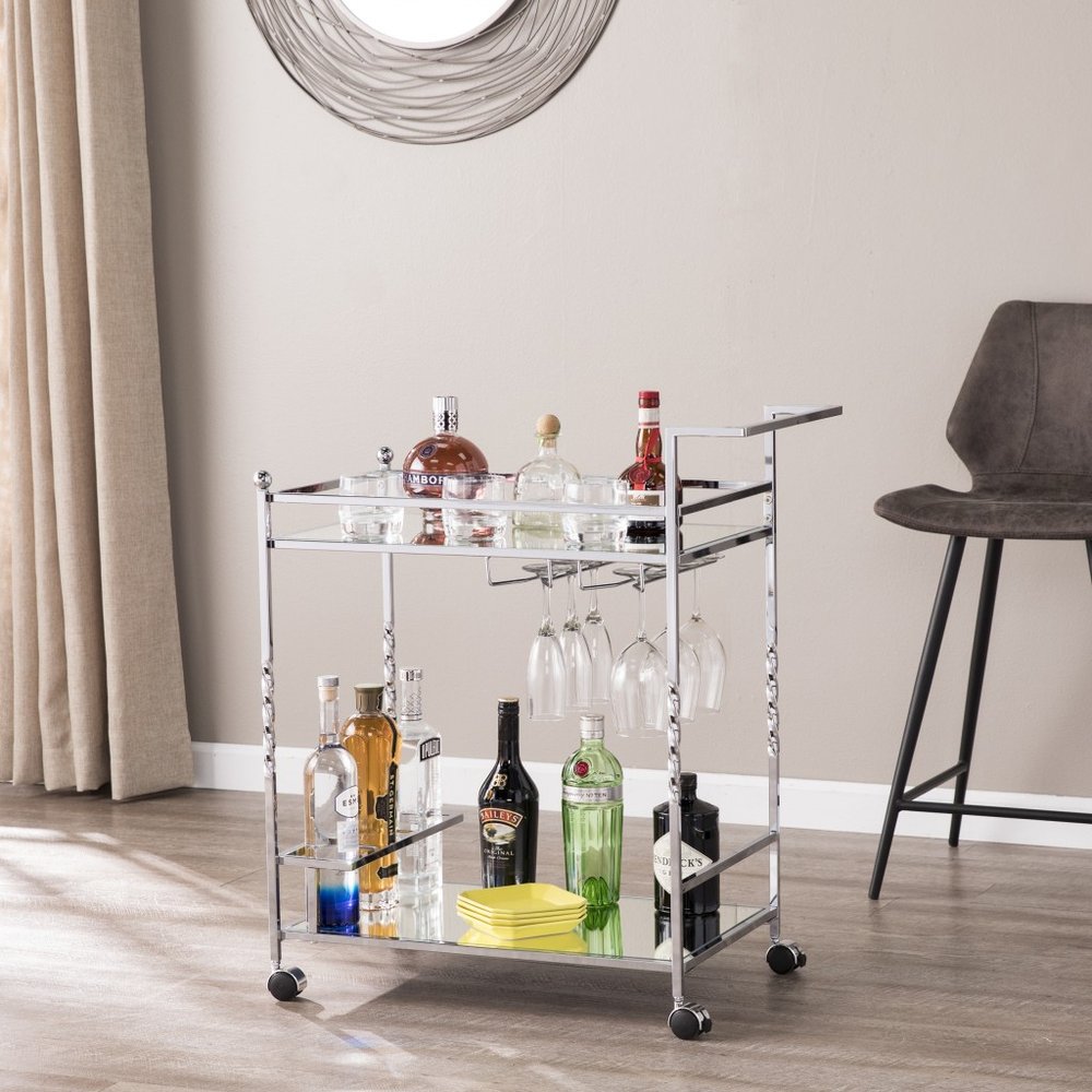 Silver metal and mirrored glass bar cart with bottles and glassware on a light-colored wall and wood floor background.