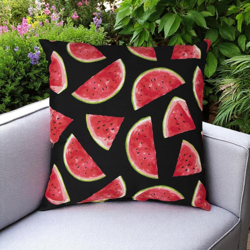 Black throw pillow with a repeating pattern of red watermelon slices, placed on a grey outdoor chair with greenery in the background.