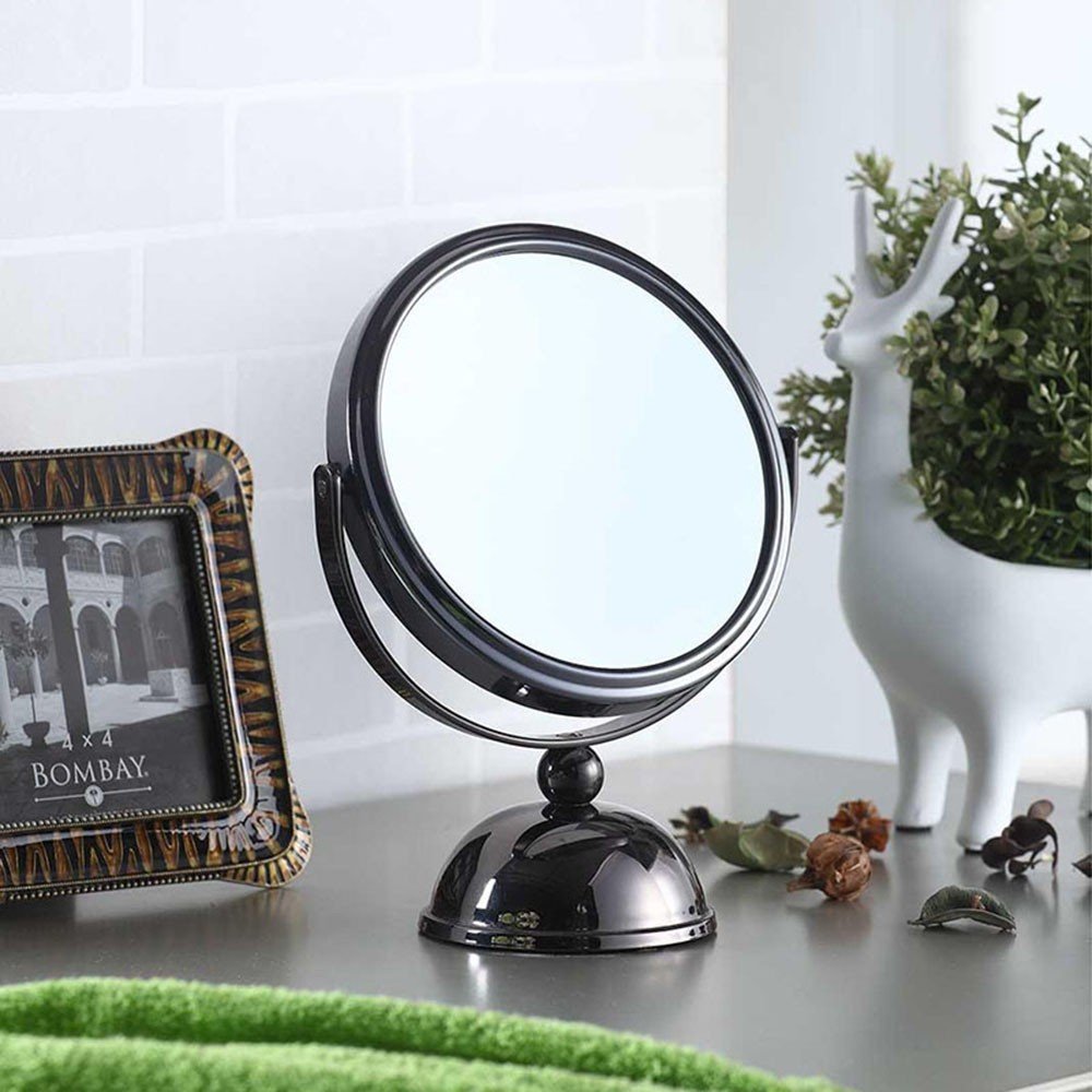 Black metal tabletop makeup mirror with a round reflective surface on a domed base, placed on a counter with a photo frame and a deer planter.