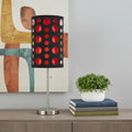 33" Steel Mod Table Lamp With Black And Red Drum Shade displayed on a dark wood tabletop in a furnished interior against a light gray wall.