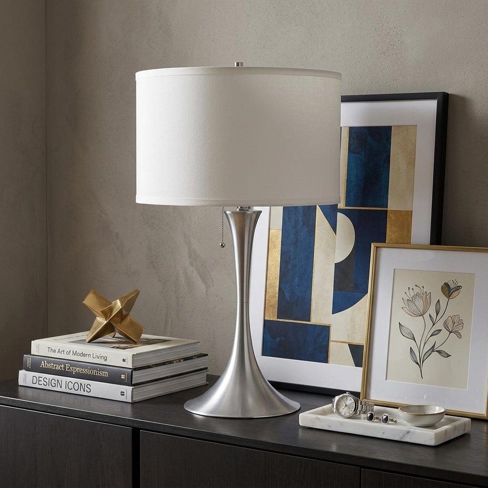 28" Silver Metal Bedside Table Lamp With White Shade displayed on a tabletop in a furnished interior against a tan wall.
